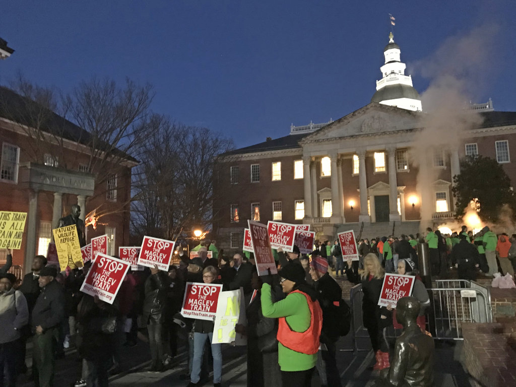 A crowd of people stands on Lawyers Mall in front of the Annapolis State House. Some wear green shirts; others carry red signs reading "Stop the Maglev Train."
