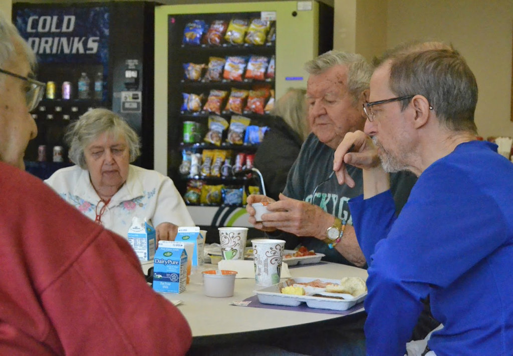 Four people sit at a round table with lunch trays and beverages in front of them. Behind them are two vending machines.