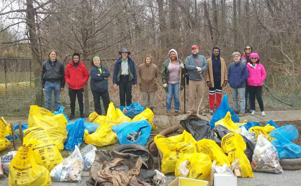 Ten people stand outside in front of a forest. In front of them is a large pile of full plastic trash bags in yellow, blue, brown and white.