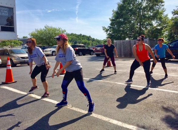 Five people wearing exercise clothes stand on pavement, feet wide apart, moving their arms back and forth in an exercise step, on a sunny day.