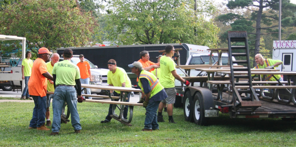 People in bright orange or yellow T-shirts lift picnic tables off a flatbed trailer and put them on the lawn.