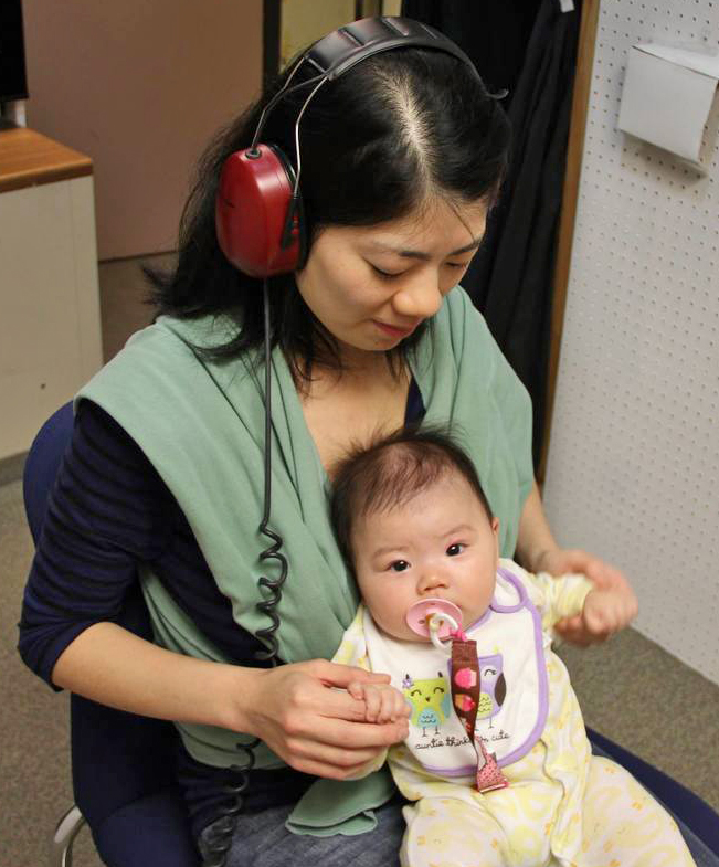 A dark-haired woman wearing large red headphones and a green shawl holds a baby on her lap. The woman looks down at the baby. The baby looks toward the viewer.