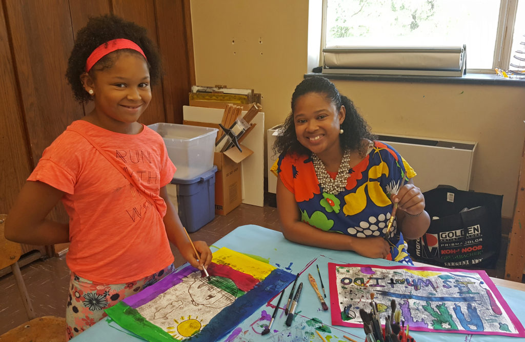 A woman in a bright floral pattern shirt sits at a table. A girl in an orange T-shirt stands next to her. Both have art supplies in front of them.