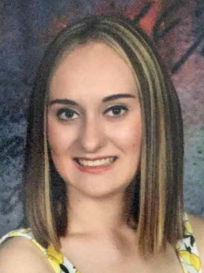 A young woman with shoulder-length blonde hair poses for the camera with a smile. She is wearing a patterned shirt of white, yellow and black.