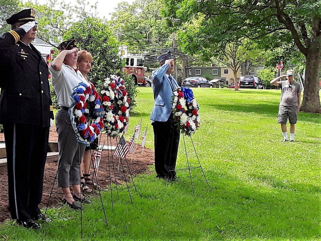 Four people stand, saluting, next to three wreaths in red, white and blue. Some distance away, another person stands, saluting.