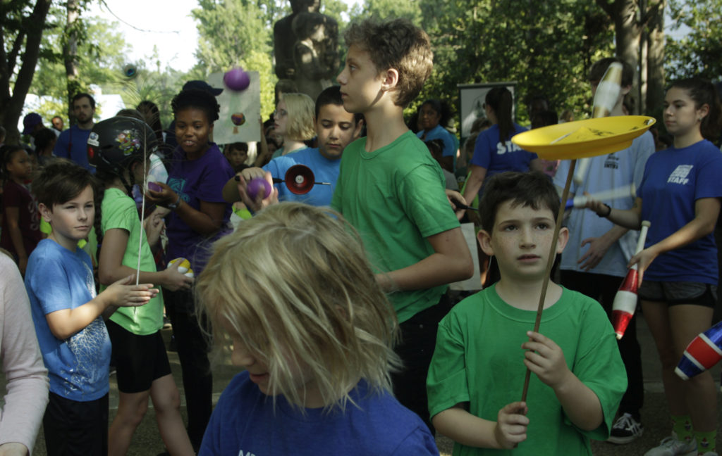 Many children stand outside in front of a statue. One child spins a yellow plate on a stick. One child is juggling balls. In the background, one child is juggling clubs.