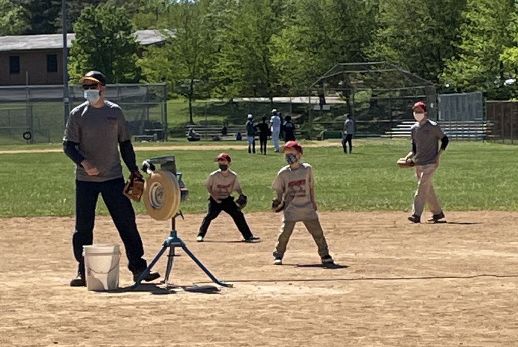 A masked adult stands next to a pitching machine on a baseball field. Nearby, two masked children in red baseball caps, wearing gloves, stand with legs apart.