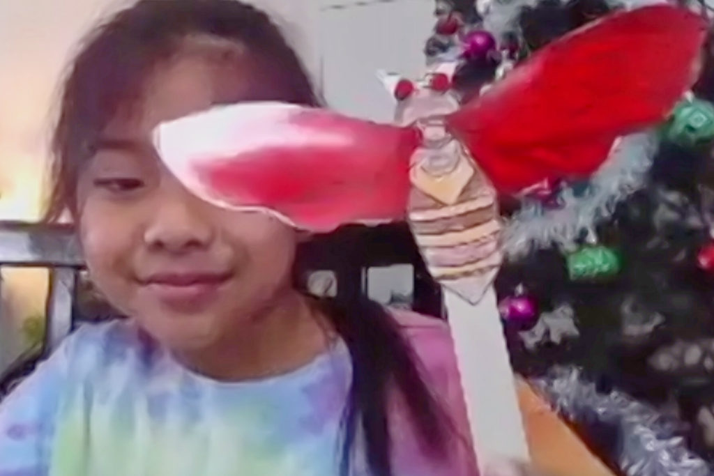 A girl with dark hair and a multicolored T-shirt holds up a puppet of a cicada: a brown bug with red eyes and big red wings. Behind her is a Christmas tree.