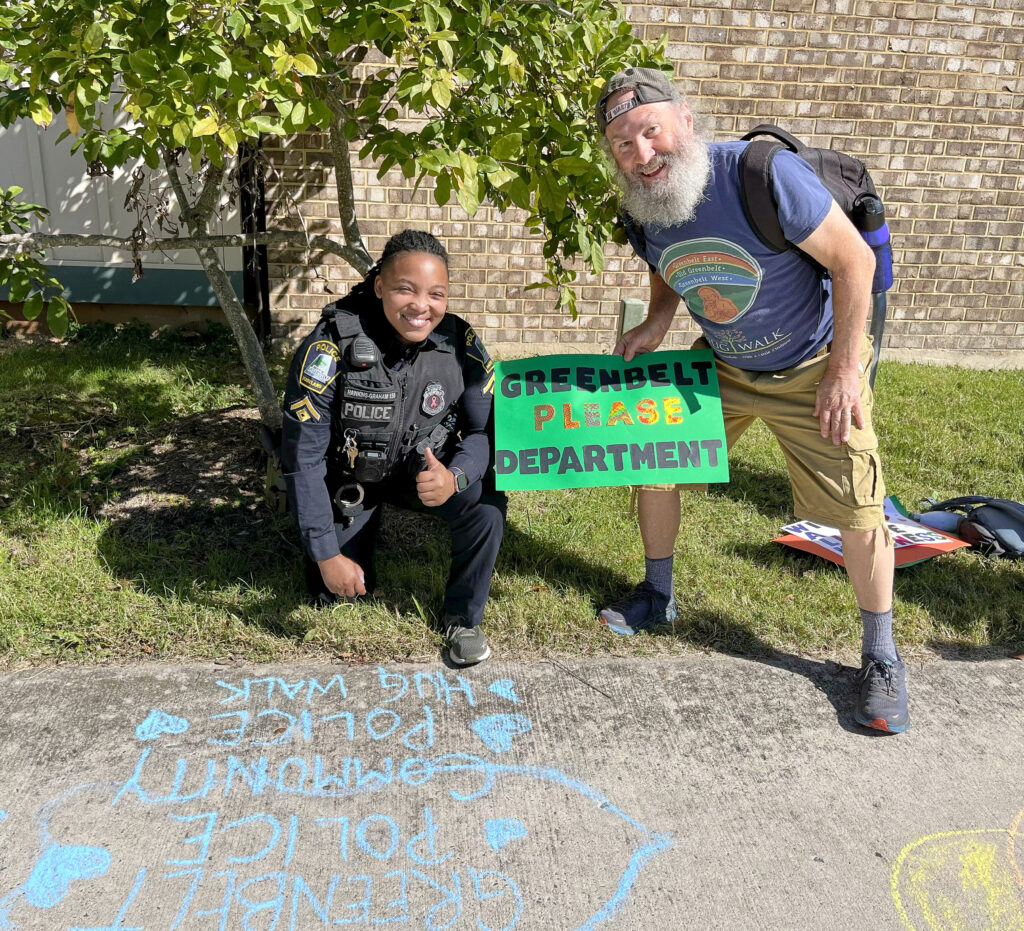 A white man with a gray beard, wearing a blue T-shirt and tan shorts, stands, crouching, next to a Black woman wearing a police uniform, who is kneeling on the ground and giving a thumbs-up. Both smile. Between them is a green sign reading "Greenbelt Please Department". On the sidewalk in front of them, blue chalk reads "Greenbelt Police, Community Police, Hug Walk."