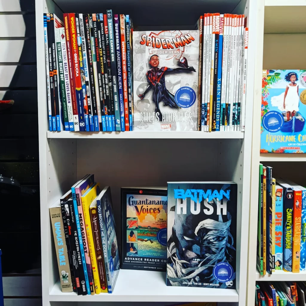 Brightly-colored books on two white bookshelves. Visible titles include Spider-Man: Spider-Verse - Miles Morales; Batman: Hush; Guantanamo Voices; and Umma's Table.