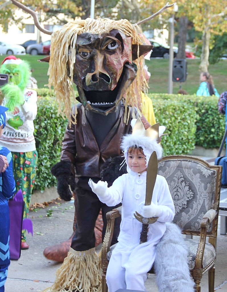A child wearing a white furry costume and a golden crown sits in an upholstered armchair with wooden arms and legs. Behind her, a standing adult wears a brown monster costume with a full-face mask.