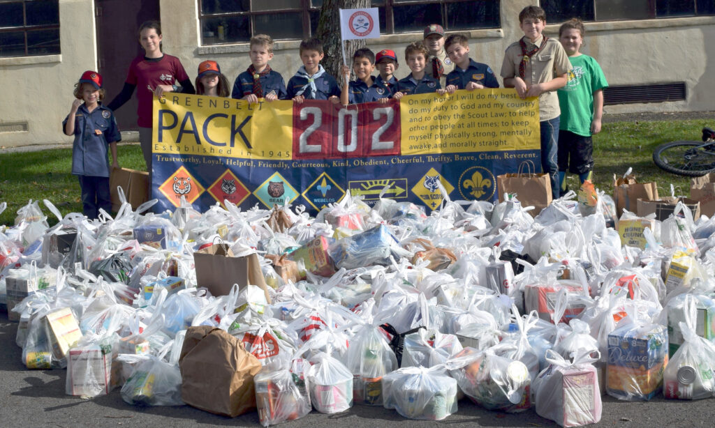 Children hold a sign saying "Greenbelt Pack 202" with Scouting logos and the Scouting pledge, standing behind a large collection of white plastic grocery bags full of food.