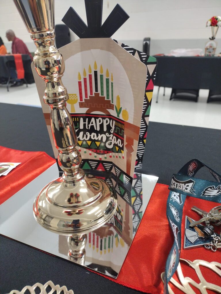 A shiny silvery candlestick sits upon a mirror. Behind it, a card reads "Happy Kwanzaa" and depicts a kinara: a branched candleholder with red, green and black candles.