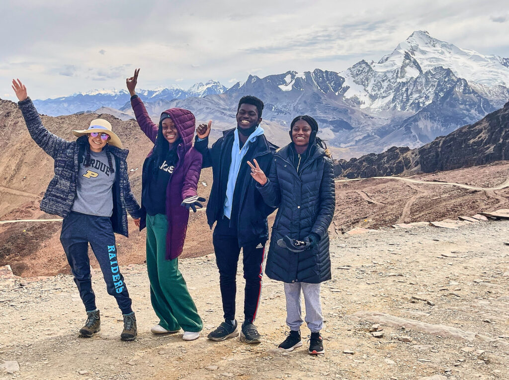 Four students stand on a plateau: two with arms extended diagonally, doing "jazz hands"; one giving a thumbs-up; and one giving a peace sign. Behind them are mountains in the distance.
