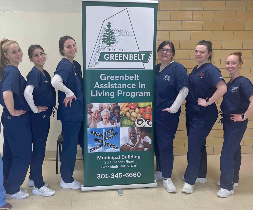 Six fair-skinned women wearing blue scrubs stand on either side of a tall sign with the Greenbelt city logo and the words "Greenbelt Assistance in Living Program."