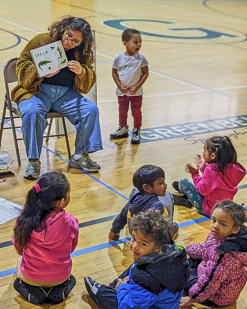 An adult sits on a chair in a gymnasium, holding a picture book open. A child stands next to the chair. Five more children sit gathered before them.