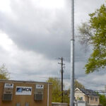 A gray monopole towers into the air, appearing roughly twice as tall as a nearby utility pole. At the top of the monopole is a triangular apparatus.