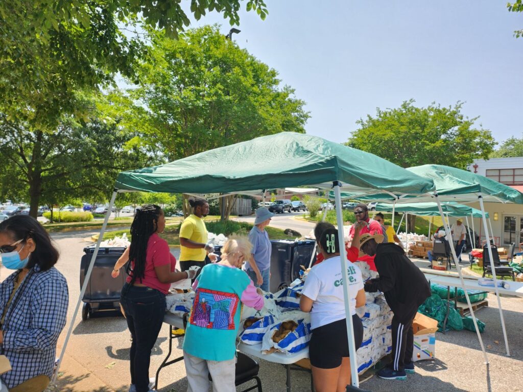 People stand below a green canopy, setting bags of food onto white plastic tables.