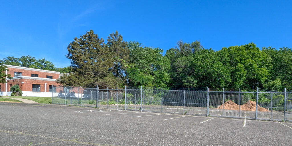 Two low piles of brown mulch, behind a chain-link fence. In the foreground, a parking lot. In the background, verdant green trees. At left, a brown brick building. A bright blue sky.