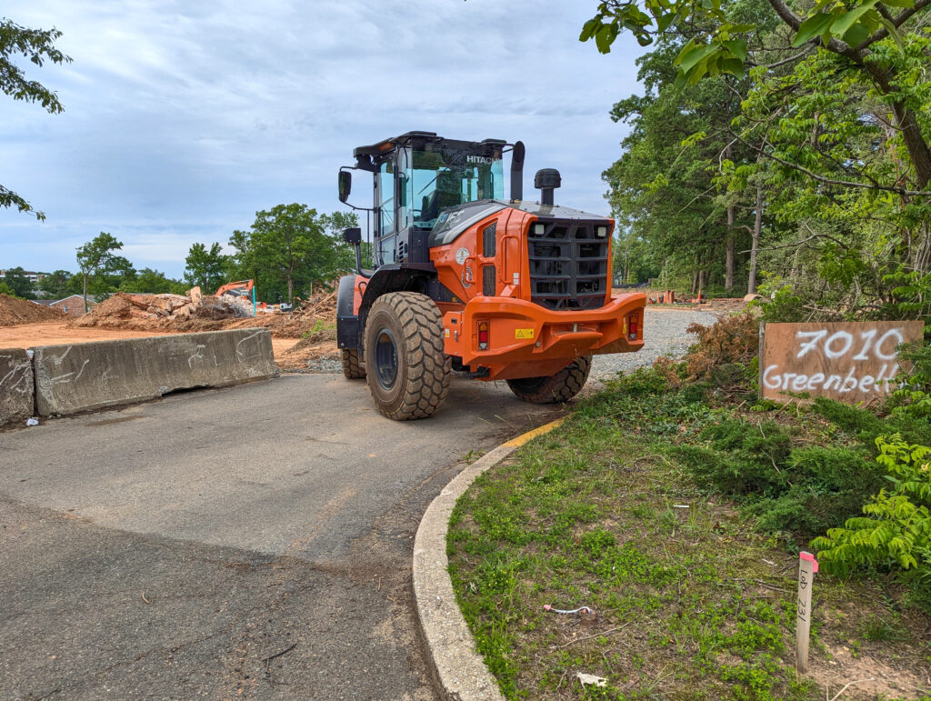 An orange construction vehicle on pavement. At left, dug-up brown dirt. At right, grass and green trees.