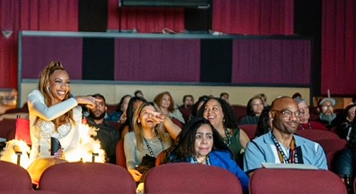 At left, A brown-skinned woman with long light-brown hair wearing a white dress. She sits in a red-and-black "director's chair" in the main auditorium of the Greenbelt Cinema. She smiles broadly and points her right index finger toward audience members sitting to her left. One audience member looks toward Lowry and points back at her, while others look ahead toward the screen.