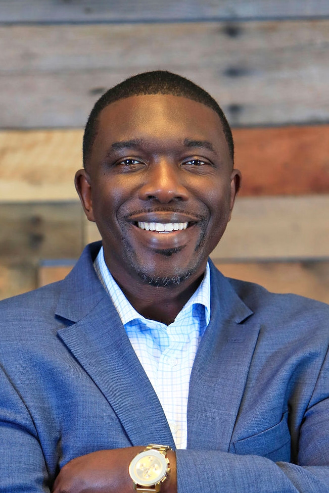A smiling brown-skinned man with short hair, wearing a blue suit and a watch.
