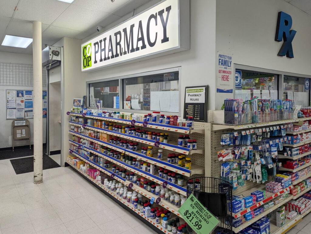 The pharmacy desk area protrudes into the pharmacy area of the co-op. A lighted sign reads "CO-OP Pharmacy" above a window and shelves of bottles of vitamins. Around the corner of the protruding area, a blue sign reads Rx, above shelves of dental hygiene supplies.