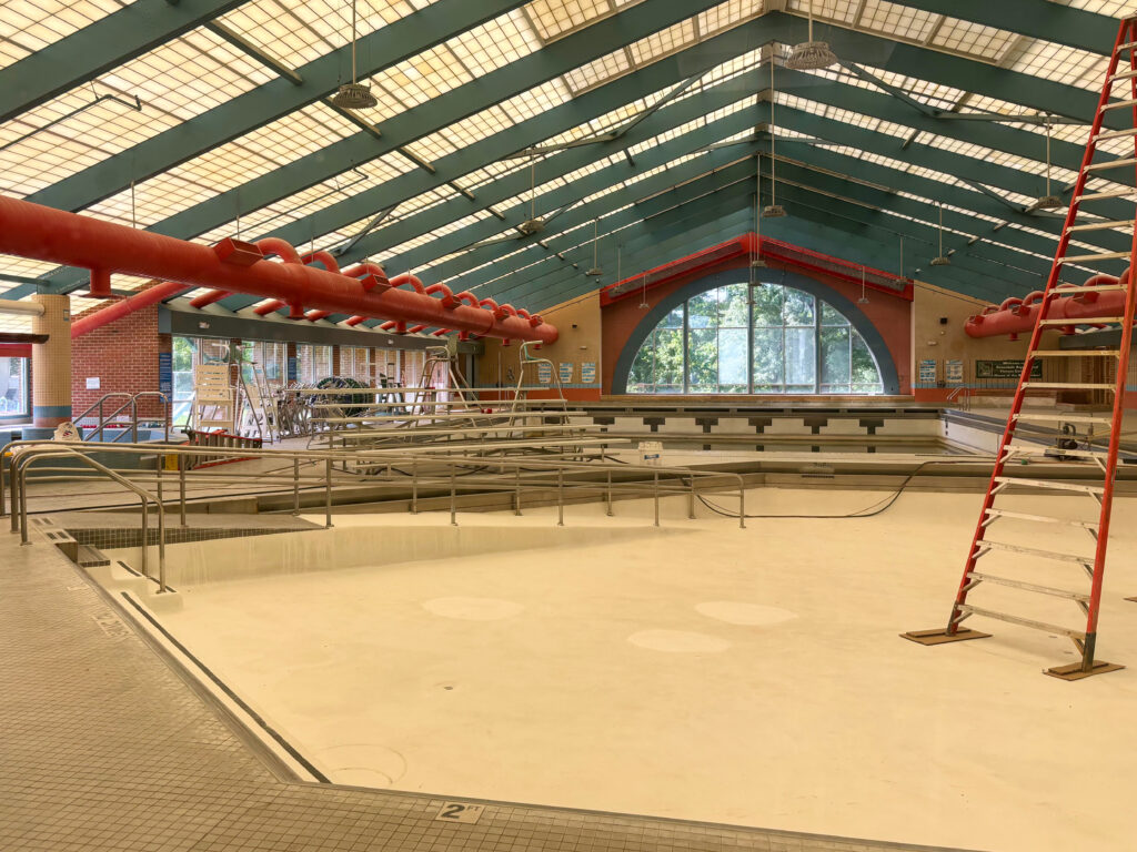 The empty indoor pool, with an orange ladder rising from its cream-colored floor. Above it, blue rafters hold up the peaked roof.