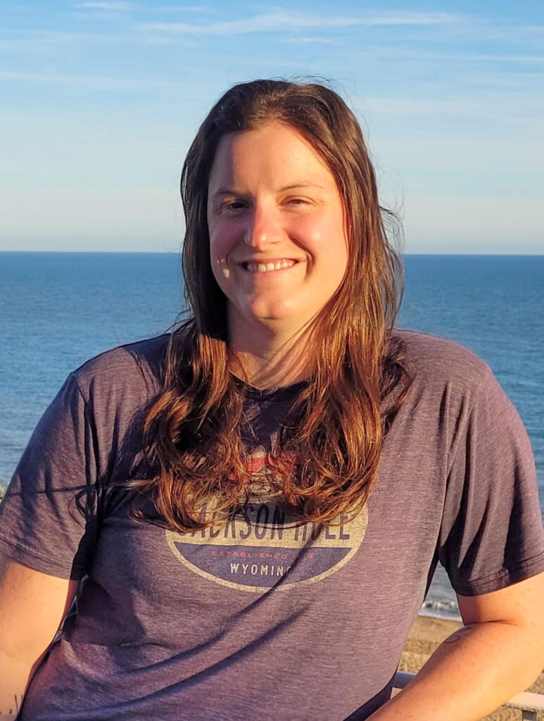 A smiling light-skinned woman with long reddish-brown hair, loose in front of her shoulders. She wears a gray T-shirt with a colorful logo. In the background, a blue body of water and a light blue sky.