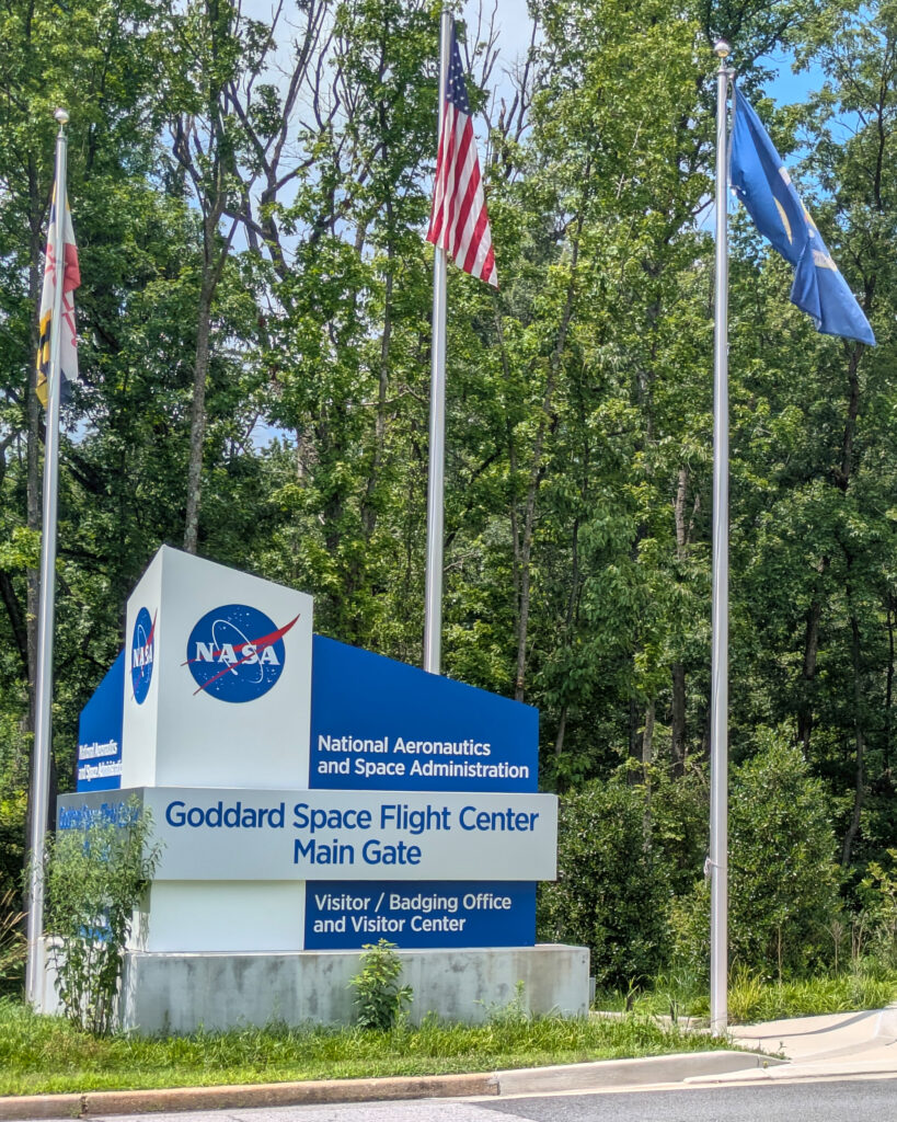 A blue and gray sign reads "National Aeronautics and Space Administration. Goddard Space Flight Center Main Gate. Visitor/Badging Office and Visitor Center", with the circular blue NASA logo. Behind the sign are three flagpoles, bearing the United States flag, the Maryland flag, and a blue flag with the NASA seal. Behind the flags are trees.