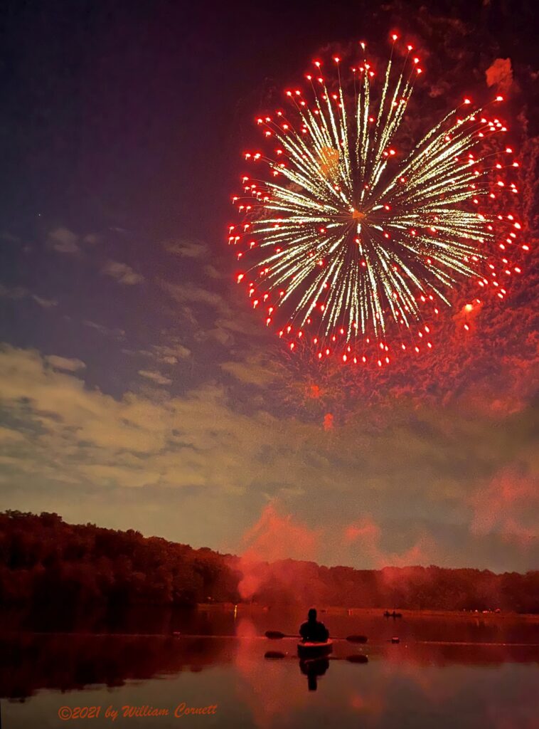 A round red and white firework explodes in the night sky above the lake. Amid clouds of red smoke just above the lake, the dark silhouette of a person in a small boat can be seen.