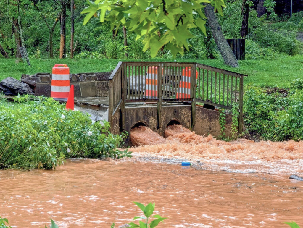 The forebay, a brown bridge with railing, connects two green areas. Behind it are orange caution barrels. A flood of brown water flows beneath it.
