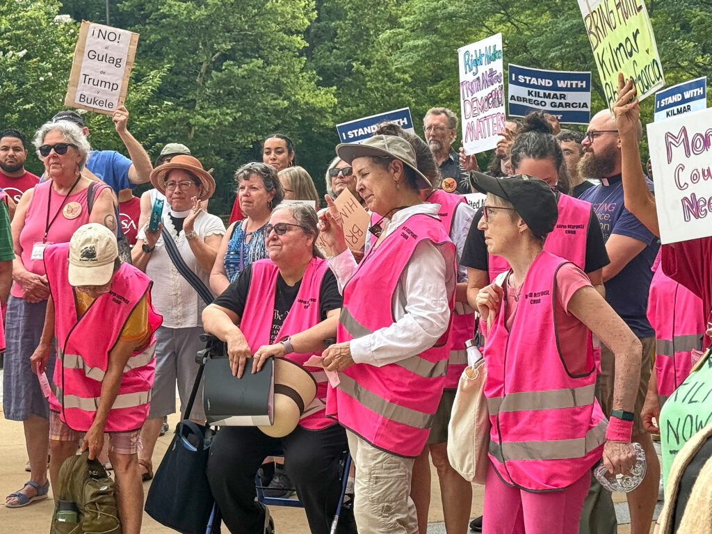 A crowd of people wearing pink vests with reflective tape. They hold signs.