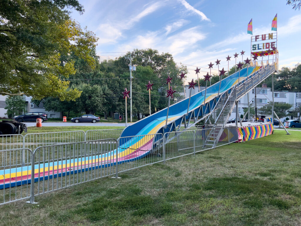 The colorful, wavy "Fun Slide" ride is set up on the grass, with trees behind it and a cloudy blue sky overhead.