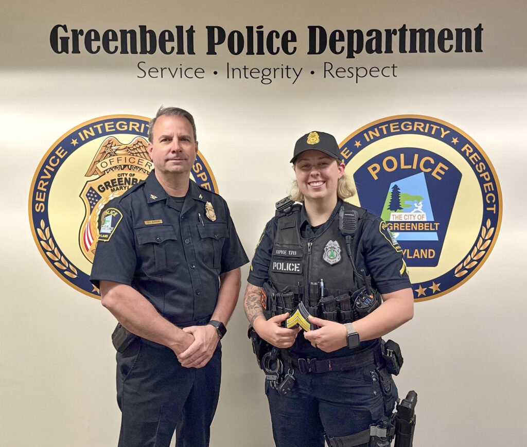 A pale-skinned dark-haired man and a pale-skinned blonde woman stand side by side. Both wear black police uniforms. The woman holds a yellow chevron award. Her uniform includes a vest and hat, which the man does not wear. The wall behind them reads "Greenbelt Police Department" and "Service, Integrity, Respect" with two round logos with blue and gold borders.