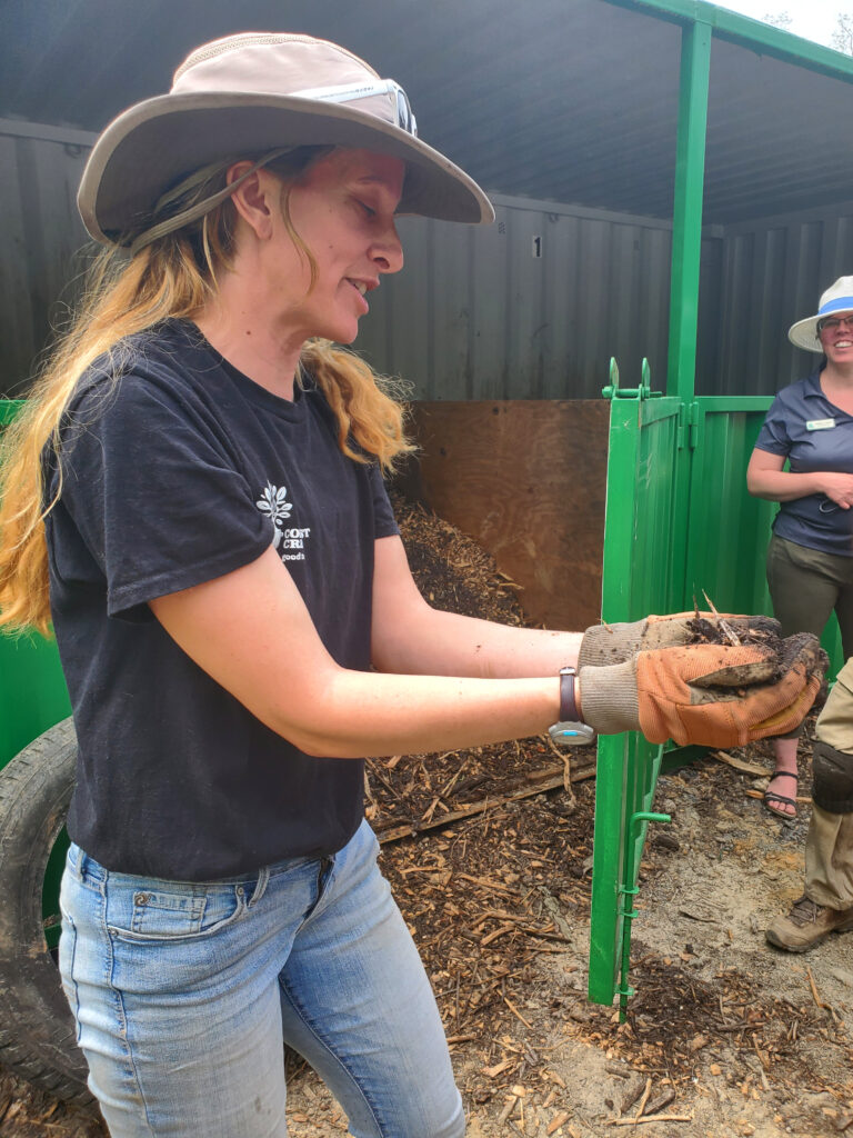 Side-profile picture of a woman with long dark-blond hair. She wears a beige wide-brimmed hat, a black T-shirt, and jeans. She wears heavy brown work gloves. In her cupped hands is a small pile of brown dirt (compost). She stands in front of a green-and-gray shed that contains a large pile of compost.