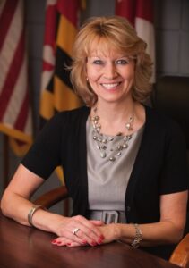 Silke Pope, a blonde woman, seated at a desk, smiling. She wears a black open jacket over a silver-gray top and gold and silver beads. The Maryland and US flags are in the background.