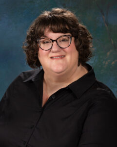 Jenni Pompi, a white woman with short brown hair and dark-framed glasses, wears a black button-down shirt.