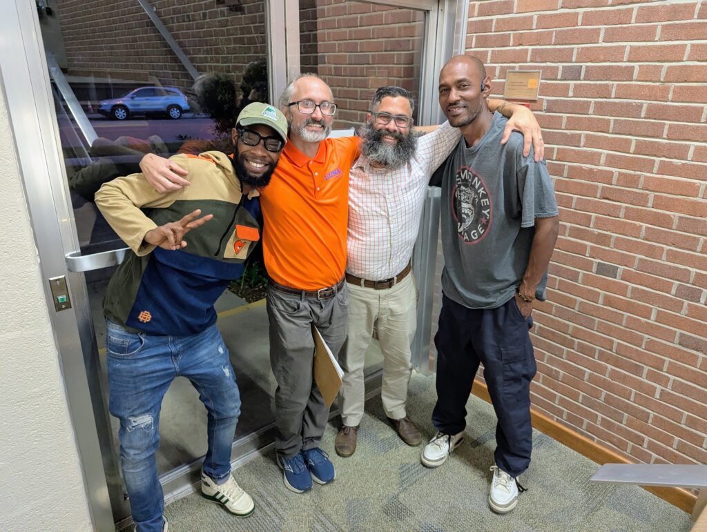 Four smiling men stand with arms around each other, in front of a glass door and a brick wall. The man at left is holding up two fingers in a V peace sign.