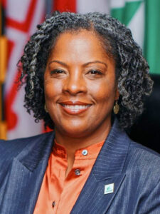 Danielle McKinney, with medium brown skin and almost-chin-length dark hair. She wars an orange blouse and blue jacket. She is in front of the Maryland flag and Greenbelt flag.