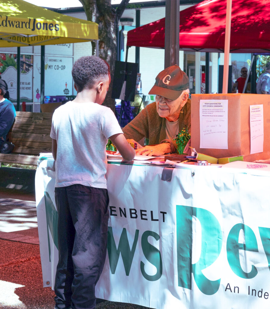 A child stands in front of an outdoor table that has the News Review logo banner. Behind the table is Mary Lou, seated, wearing a dark baseball cap. In the background are red and yellow canopies.