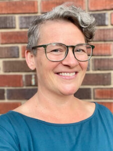 Amy Knesel, a white woman with short gray hair and glasses, wears a blue top and long earrings. Photo courtesy of Amy Knesel. 