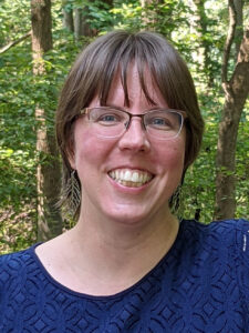 Kristen Weaver, a white woman with light brown hair in bangs, smiles directly at the camera. She is wearing a blue top and light-framed glasses.