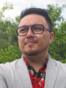 Frankie Fritz, a young man with a slight goatee and moustache and dark brown hair, looks slightly to the right. He is wearing a patterned red and black shirt under a light gray blazer, and dark-framed glasses.