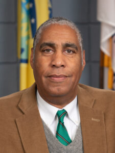 Emmett Jordan, with medium brown skin and close-cut gray hair, looks directly into the camera. He is wearing a camel brown blazer over a white shirt, green striped tie and gray sweater. There are flags in the background.