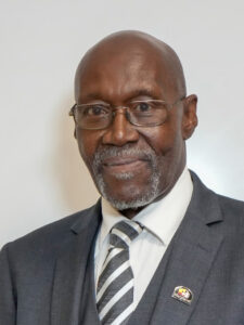 Kevin Lockhart (Coach K), an older Black man with a gray mustache and goatee, wears a gray suit, white shirt, and gray and white tie, with a Maryland pin on his lapel.