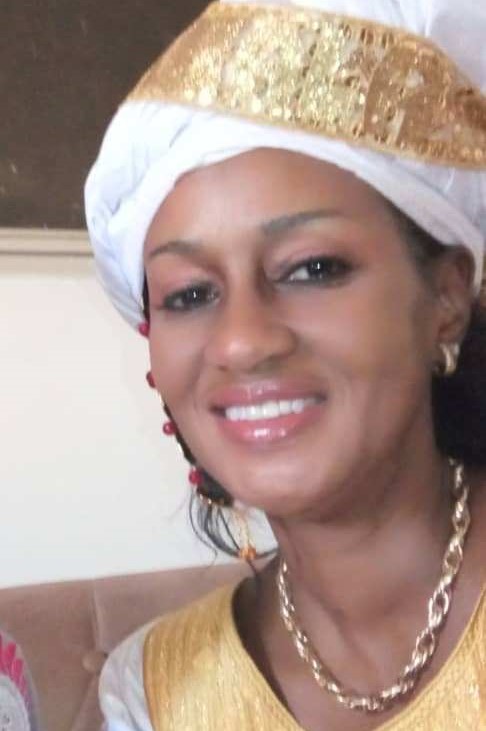 A smiling woman with medium brown skin, wearing a gold-and-white blouse and headwrap, and a necklace.