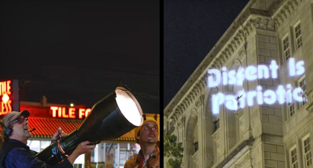 A night scene, with a split image. In the left half, a man at left holds a conical lighting device. Another man stands to the right, looking toward the right. Behind them is a building with red neon lights spelling out TILE FOR LESS. In the right half of the split image, the words "Dissent is Patriotic" in white light are projected on a light beige building.
