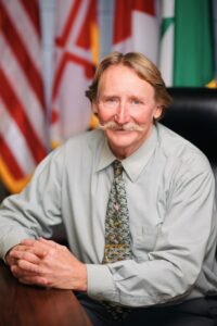 Rodney Roberts, a white man with sandy hair and a large mustache, sits at a table wearing a pale green shirt and patterned tie. Flags are in the background.