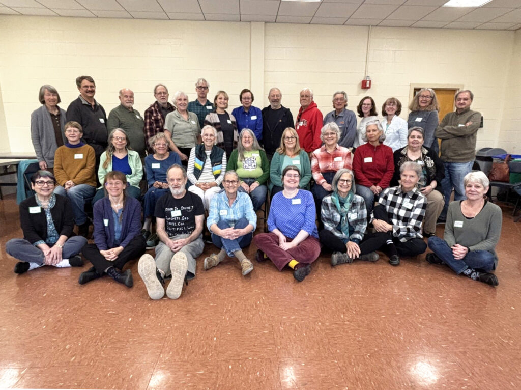 People pose for a photo in three rows: the back row standing, the middle row seated on chairs, and the front row sitting on the floor.